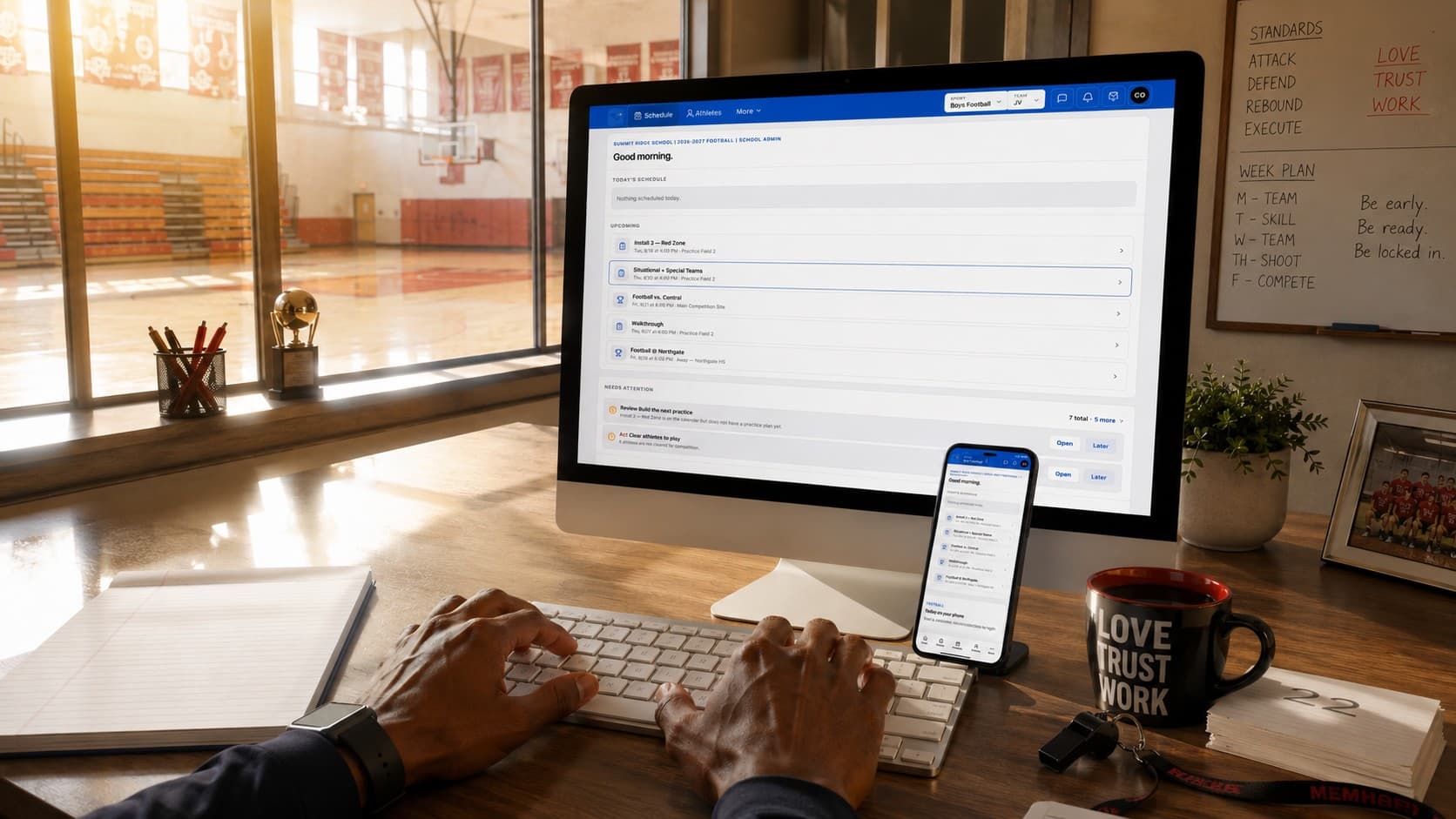 First-person view of a coach's desk: The Program Suite home dashboard on the monitor, the coach app on an iPhone, the gym through the window